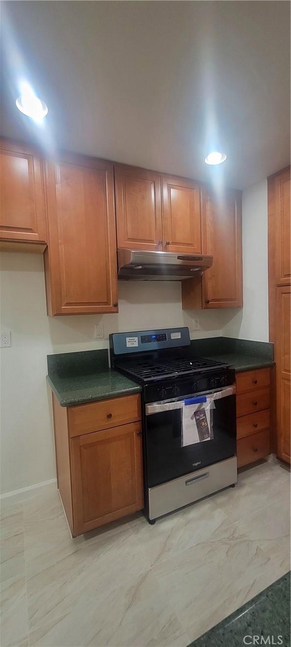 kitchen featuring dark countertops, baseboards, under cabinet range hood, brown cabinets, and gas stove