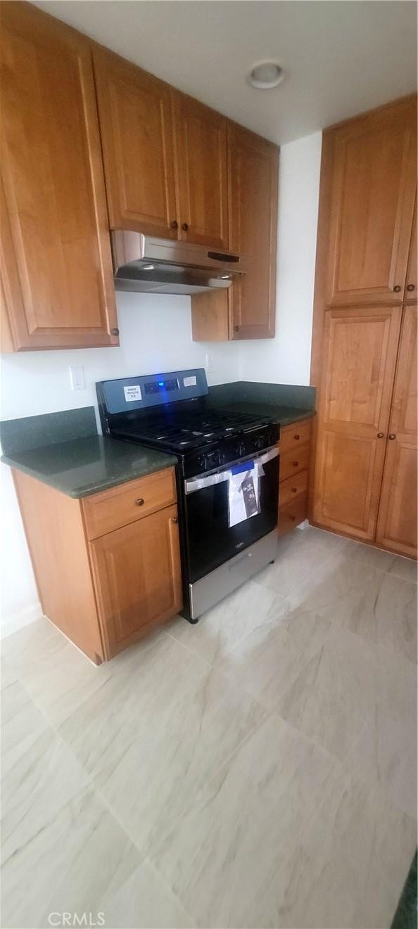 kitchen featuring stainless steel range with gas stovetop, dark countertops, under cabinet range hood, and brown cabinetry