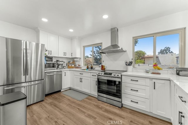 kitchen with wall chimney range hood, white cabinetry, a sink, stainless steel appliances, and light wood-style flooring