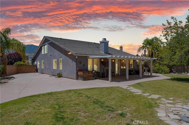 back of house at dusk with a yard, fence, a chimney, and a patio area