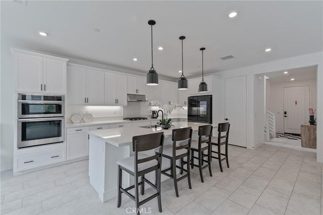 kitchen with under cabinet range hood, stainless steel double oven, white cabinetry, and an island with sink