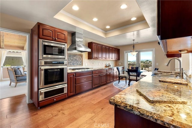 kitchen featuring a sink, a raised ceiling, light wood finished floors, wall chimney exhaust hood, and stainless steel appliances