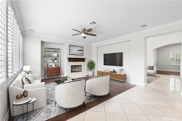 living room featuring crown molding, arched walkways, a tiled fireplace, a ceiling fan, and tile patterned flooring