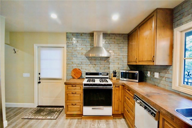 kitchen featuring wall chimney range hood, backsplash, brown cabinets, light wood-style floors, and white appliances