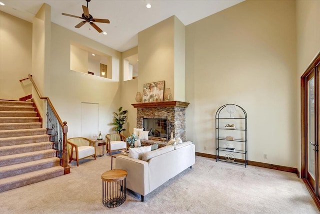 carpeted living area featuring a stone fireplace, a ceiling fan, baseboards, a towering ceiling, and stairway