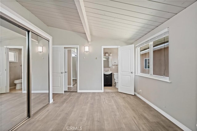 unfurnished bedroom featuring a closet, light wood-style floors, baseboards, ensuite bathroom, and lofted ceiling with beams