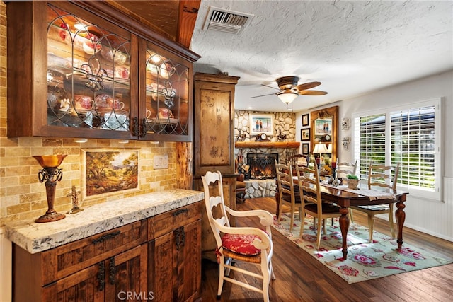dining space featuring dark wood finished floors, visible vents, a textured ceiling, a stone fireplace, and ceiling fan