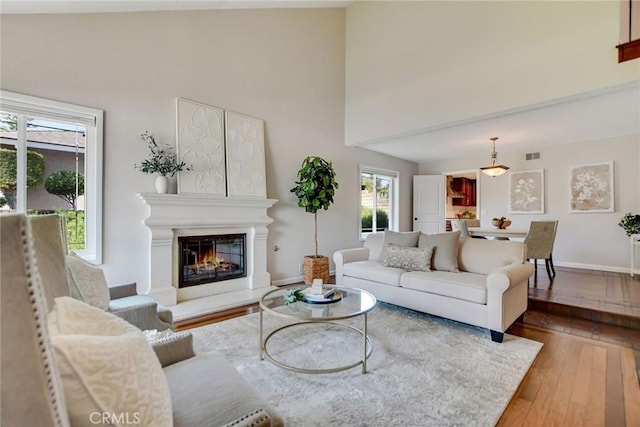 living room featuring visible vents, wood-type flooring, a glass covered fireplace, baseboards, and high vaulted ceiling