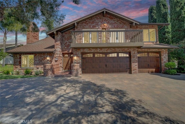 view of front of house with aphalt driveway, a balcony, a chimney, brick siding, and an attached garage
