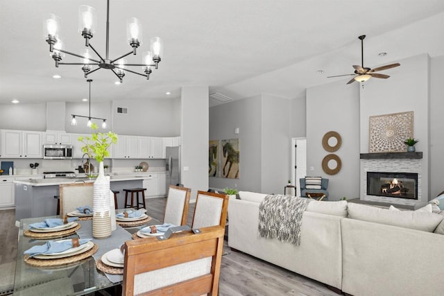 dining room featuring visible vents, high vaulted ceiling, a glass covered fireplace, ceiling fan with notable chandelier, and light wood-style floors