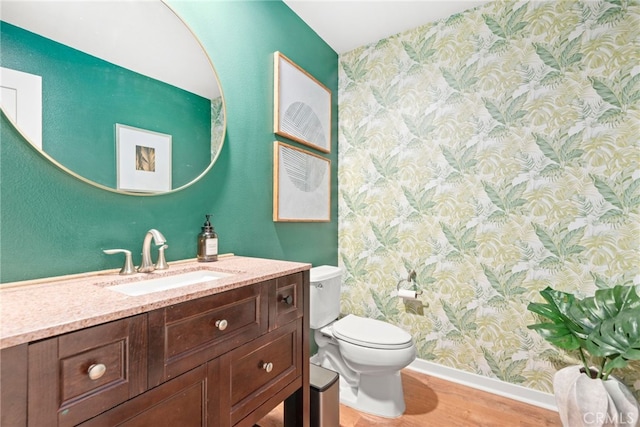 bathroom with wood finished floors, toilet, vanity, baseboards, and wallpapered walls