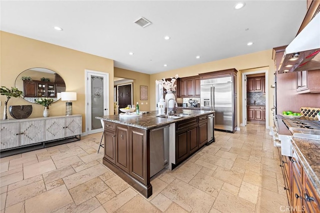 kitchen with appliances with stainless steel finishes, a sink, a kitchen island with sink, extractor fan, and stone tile floors