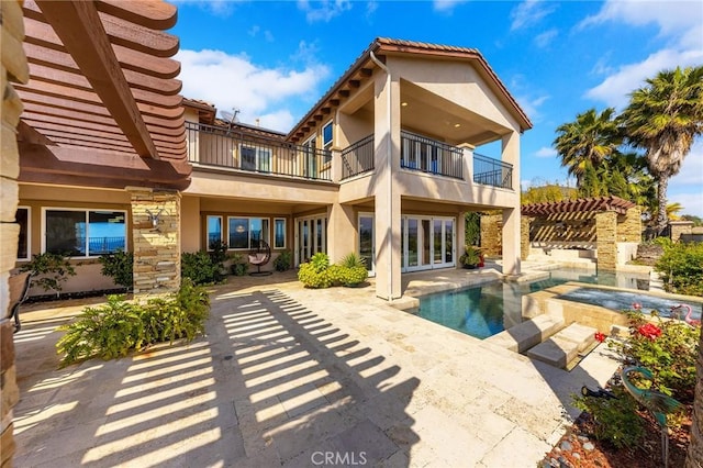 back of property featuring a balcony, stone siding, a patio, an outdoor pool, and stucco siding