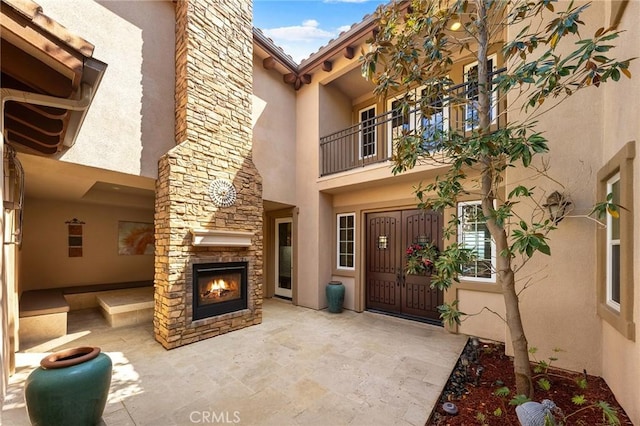 entrance to property featuring a balcony, stone siding, an outdoor stone fireplace, a patio, and stucco siding