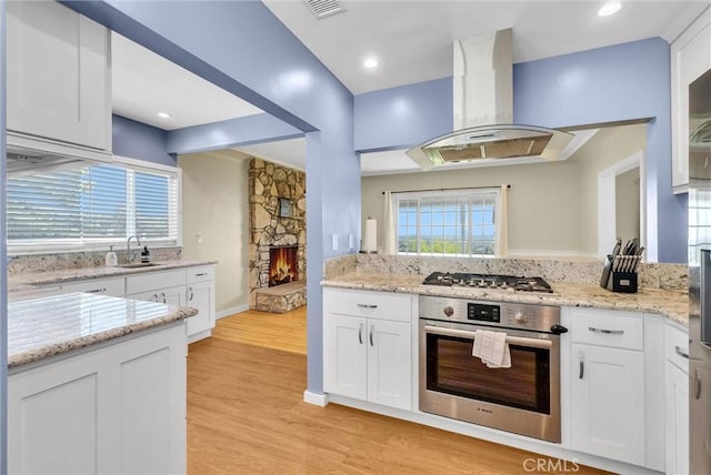 kitchen featuring appliances with stainless steel finishes, a sink, a fireplace, white cabinetry, and island range hood