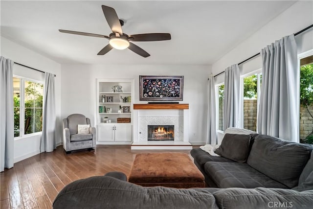 living room featuring built in shelves, a tiled fireplace, a ceiling fan, baseboards, and dark wood-style floors