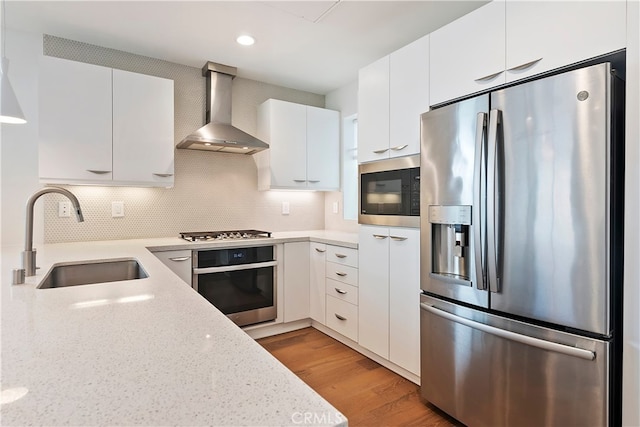 kitchen featuring wall chimney range hood, light wood-type flooring, white cabinetry, a sink, and stainless steel appliances