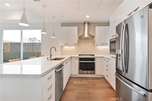 kitchen with a peninsula, wall chimney range hood, a sink, decorative light fixtures, and stainless steel appliances