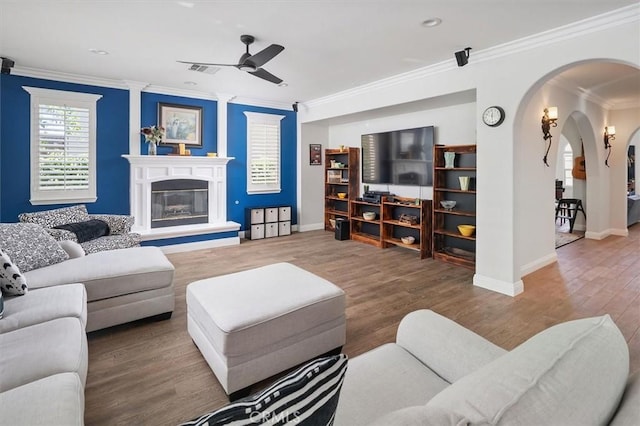living room with ornamental molding, a glass covered fireplace, wood finished floors, ceiling fan, and arched walkways