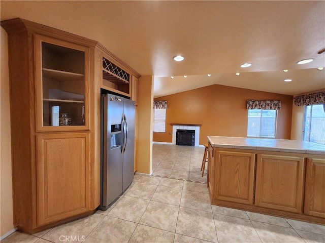 kitchen with light tile patterned floors, a fireplace, stainless steel fridge with ice dispenser, tile countertops, and vaulted ceiling