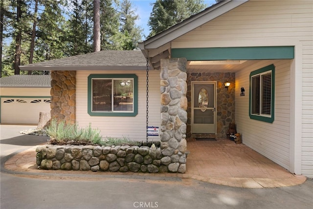 view of exterior entry with driveway, a shingled roof, stone siding, and a garage
