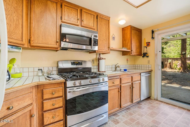 kitchen with stone finish floor, brown cabinetry, appliances with stainless steel finishes, a sink, and tile counters
