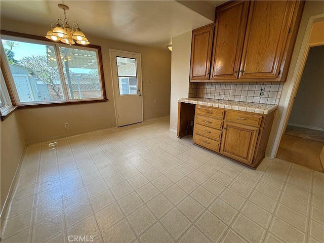 kitchen with brown cabinetry, tile countertops, backsplash, baseboards, and an inviting chandelier