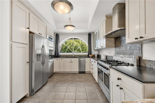 kitchen featuring wall chimney range hood, dark countertops, stainless steel appliances, tasteful backsplash, and a tray ceiling