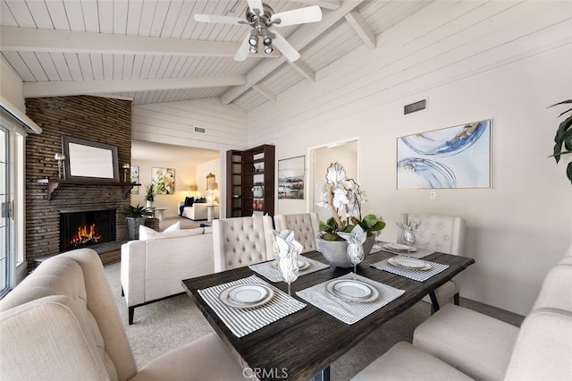carpeted dining room with ceiling fan, a stone fireplace, wooden ceiling, vaulted ceiling with beams, and visible vents