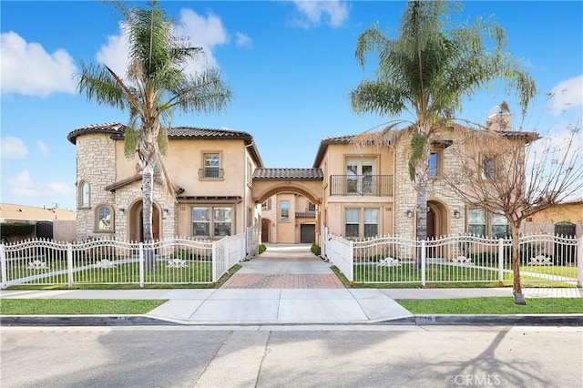 mediterranean / spanish-style home featuring a balcony, stone siding, stucco siding, a fenced front yard, and a tile roof