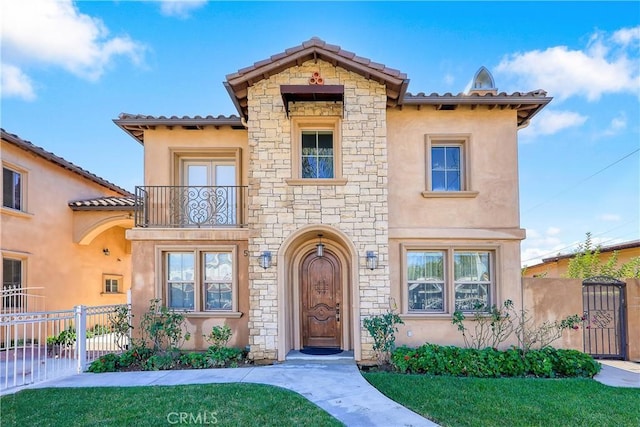 mediterranean / spanish-style house with fence, a front lawn, stone siding, stucco siding, and a balcony