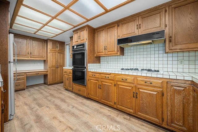 kitchen with dobule oven black, freestanding refrigerator, light wood-type flooring, under cabinet range hood, and tasteful backsplash
