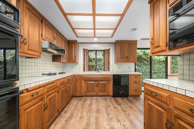 kitchen featuring brown cabinetry, tile countertops, under cabinet range hood, and black appliances