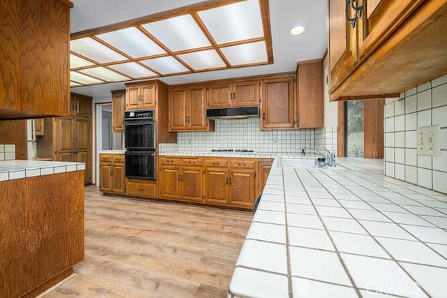 kitchen with dobule oven black, brown cabinetry, tile counters, under cabinet range hood, and tasteful backsplash