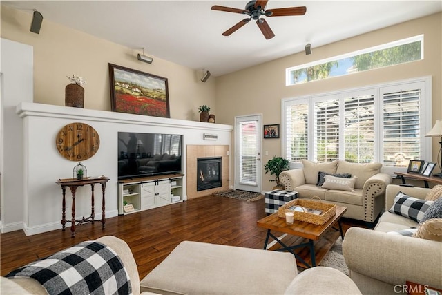 living room featuring a towering ceiling, wood finished floors, baseboards, a tile fireplace, and a ceiling fan