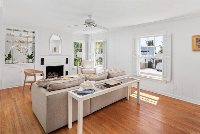 living room featuring ceiling fan, crown molding, a brick fireplace, light wood finished floors, and a decorative wall