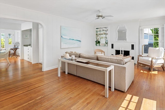 living area featuring ceiling fan, light wood finished floors, a fireplace, arched walkways, and ornamental molding