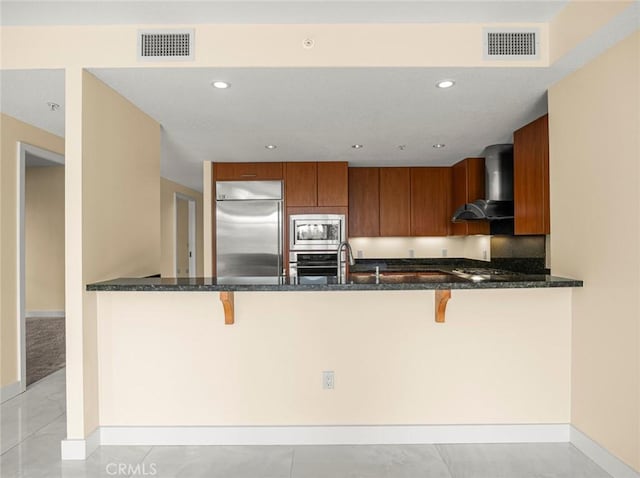 kitchen with wall chimney range hood, built in appliances, brown cabinetry, and visible vents