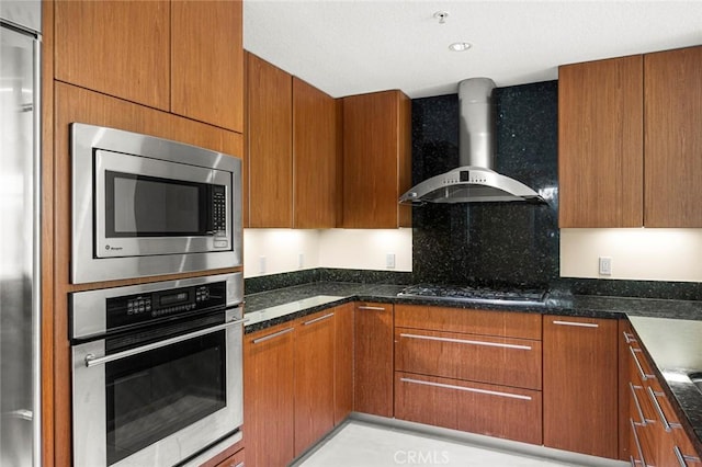 kitchen featuring built in appliances, brown cabinets, dark stone counters, and wall chimney exhaust hood