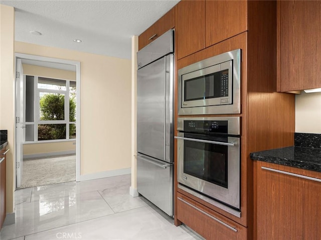 kitchen featuring brown cabinetry, a textured ceiling, baseboards, dark stone countertops, and built in appliances