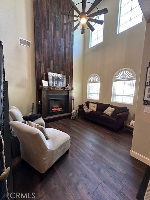 living room with ceiling fan, a high ceiling, a glass covered fireplace, visible vents, and dark wood-style floors