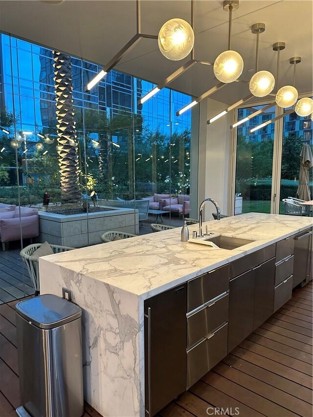 kitchen with dark wood-style flooring, hanging light fixtures, light stone counters, a sink, and a center island with sink