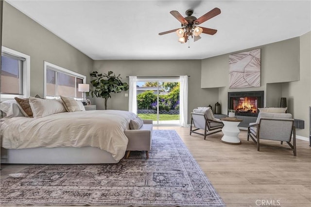 bedroom with light wood-type flooring, ceiling fan, a lit fireplace, and access to exterior