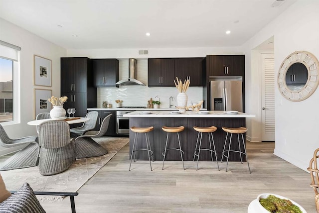kitchen with wall chimney range hood, light countertops, a kitchen breakfast bar, stainless steel appliances, and a kitchen island