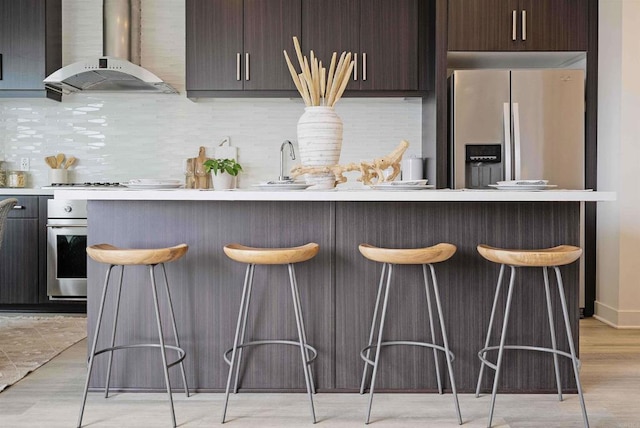 kitchen with dark brown cabinets, a kitchen breakfast bar, wall chimney range hood, tasteful backsplash, and stainless steel appliances