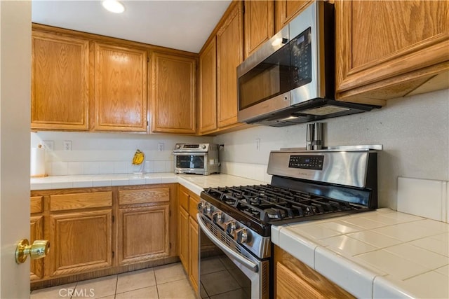 kitchen featuring brown cabinetry, appliances with stainless steel finishes, light tile patterned floors, a toaster, and tile counters