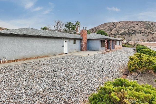 rear view of house with a shingled roof, a mountain view, stucco siding, and a patio