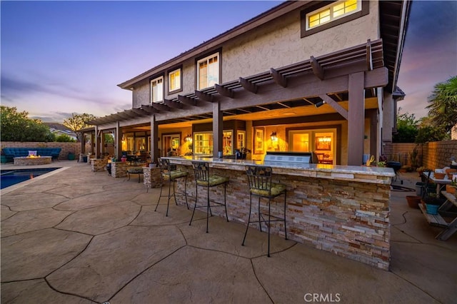 patio terrace at dusk with a pergola, fence, outdoor dry bar, and an outdoor fire pit