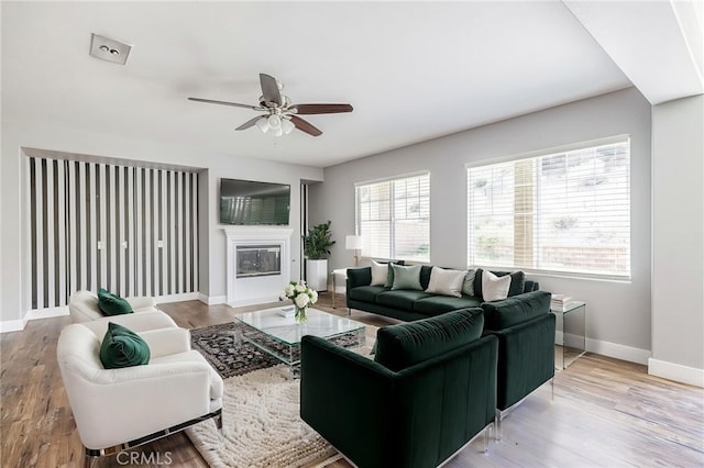 living room featuring baseboards, a glass covered fireplace, ceiling fan, and wood finished floors