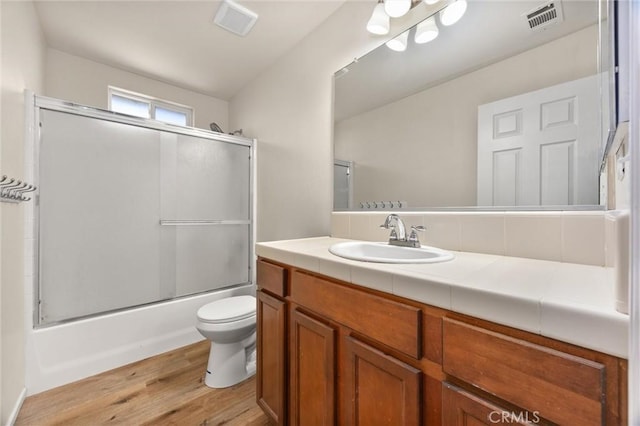 bathroom with vanity, toilet, enclosed tub / shower combo, wood finished floors, and visible vents